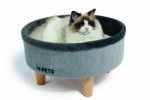 A fluffy tabby cat relaxes in a round, elevated cat bed with a soft fur lining and sleek wooden legs. The modern design complements the cat's stylish home decor.